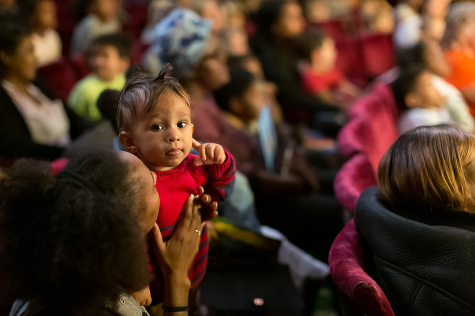 An audience sitting in a theatre watching a Tall Stories performance with focus on a young girl being help up by her mother