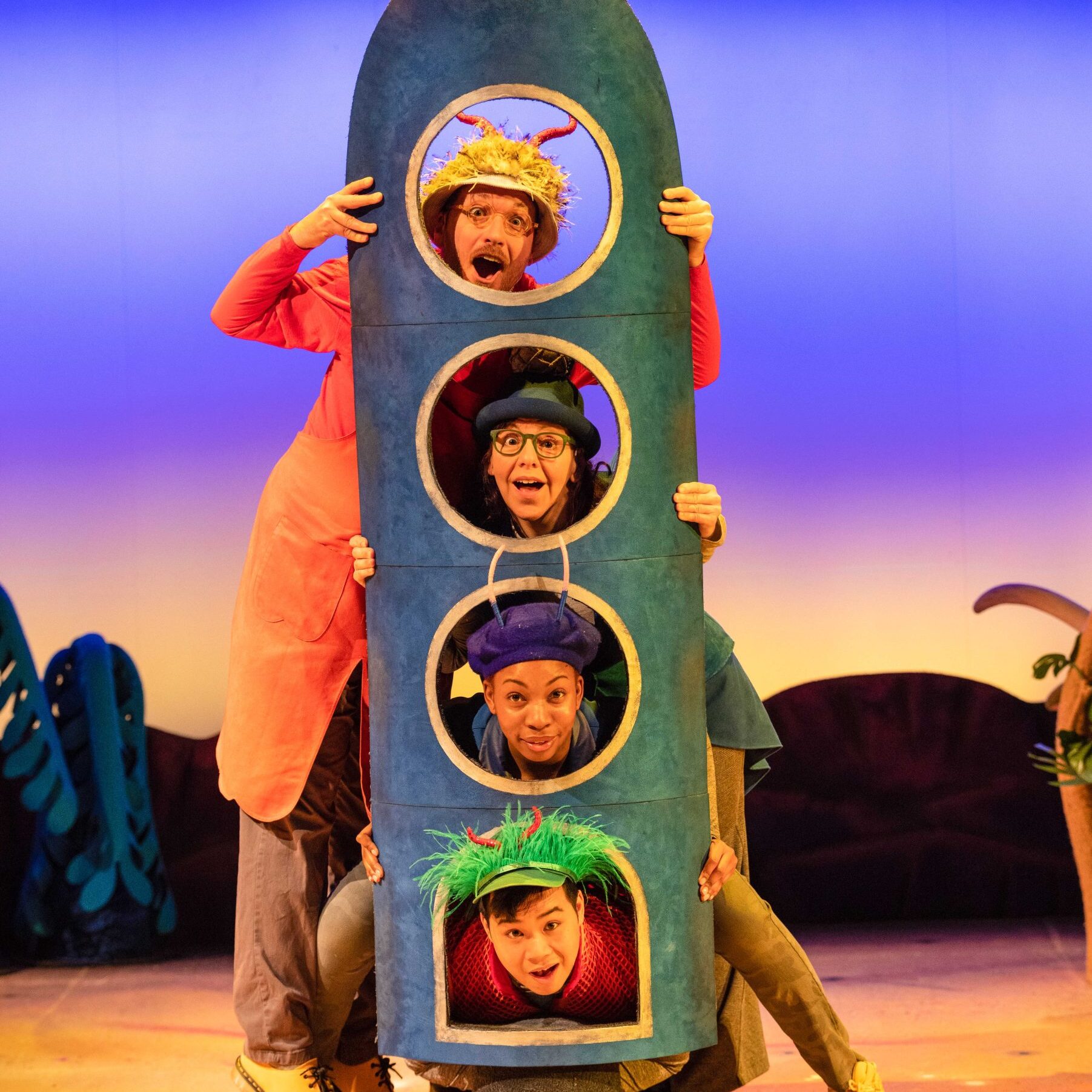 Two actors playing red Smeds and two actresses playing blue Smoos peeping therough the portholes of a rocket. The rocket is tall and thing and the portholes are one on top of the other.