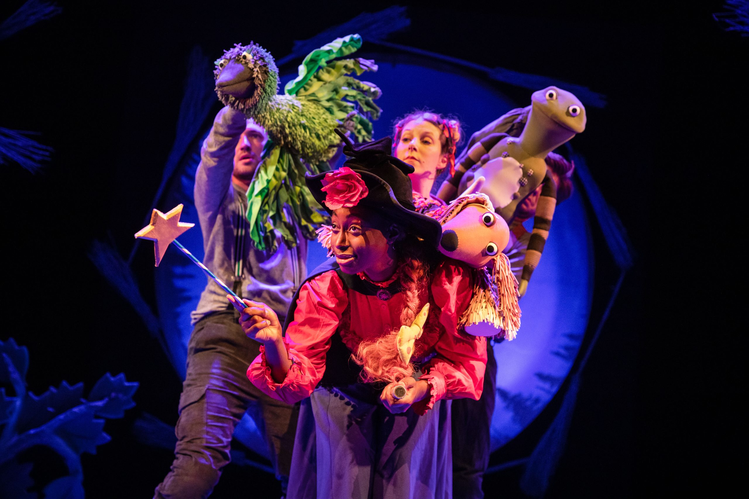 The witch wearing a black hat and a red blouse holding a magic wand with a star on the end. The hat has a pink flower on and she has a yellow spotty bow in her hair. A dog puppet is on her left shoulder and a frog puppet is peeping over him. The green bird puppet is flying over her right shoulder, controlled by a puppeteer dressed in grey. An actress with blonde hair has her head popping out above the witch's hat.
