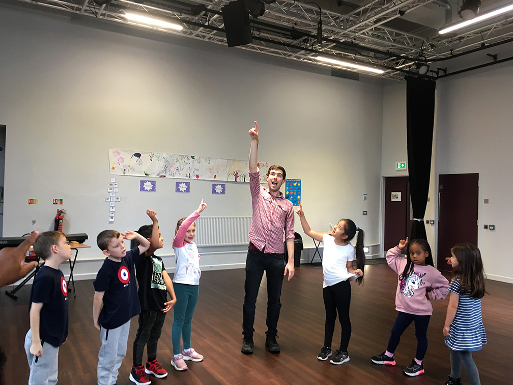 A group of children and an adult pointing up to the ceiling in the studio. the adult male is wearing a pink shirt and black trousers.