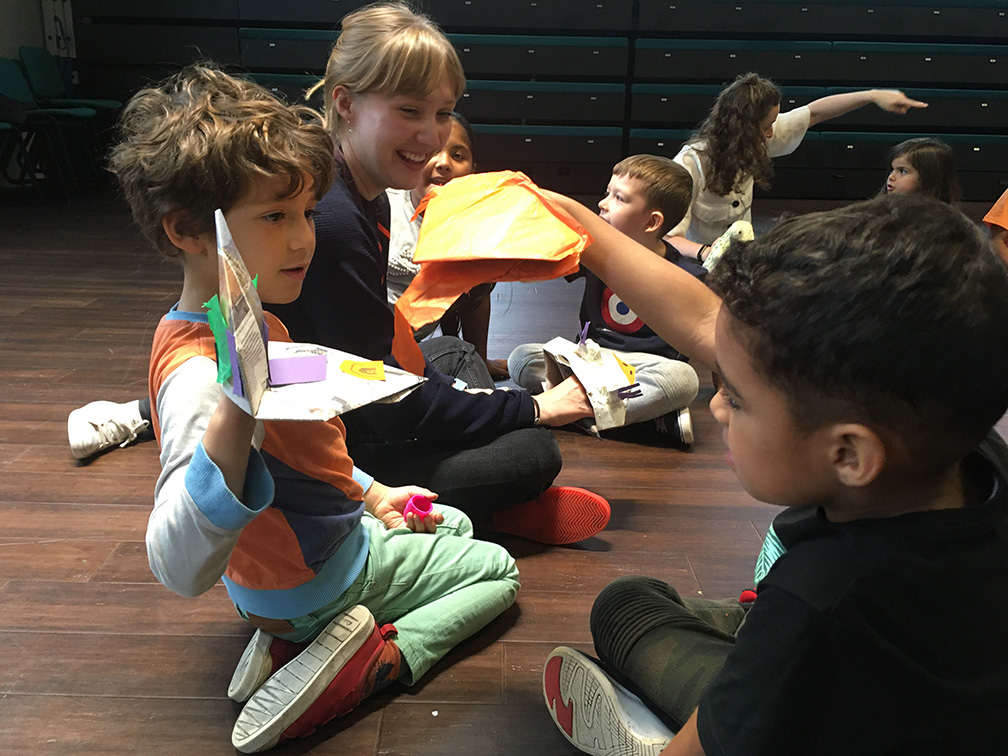 Some children playing with paper whilst an adult looks on, laughing.