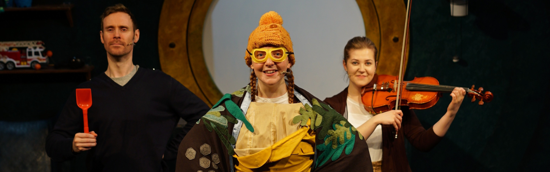 Three actors standing face on to camera. The middle one is wearing a sea themed blanket as a cape with yellow hat and goggles. The man on the left is in a navy jumper and holding a red spade. The woman on the right is playing a viola.