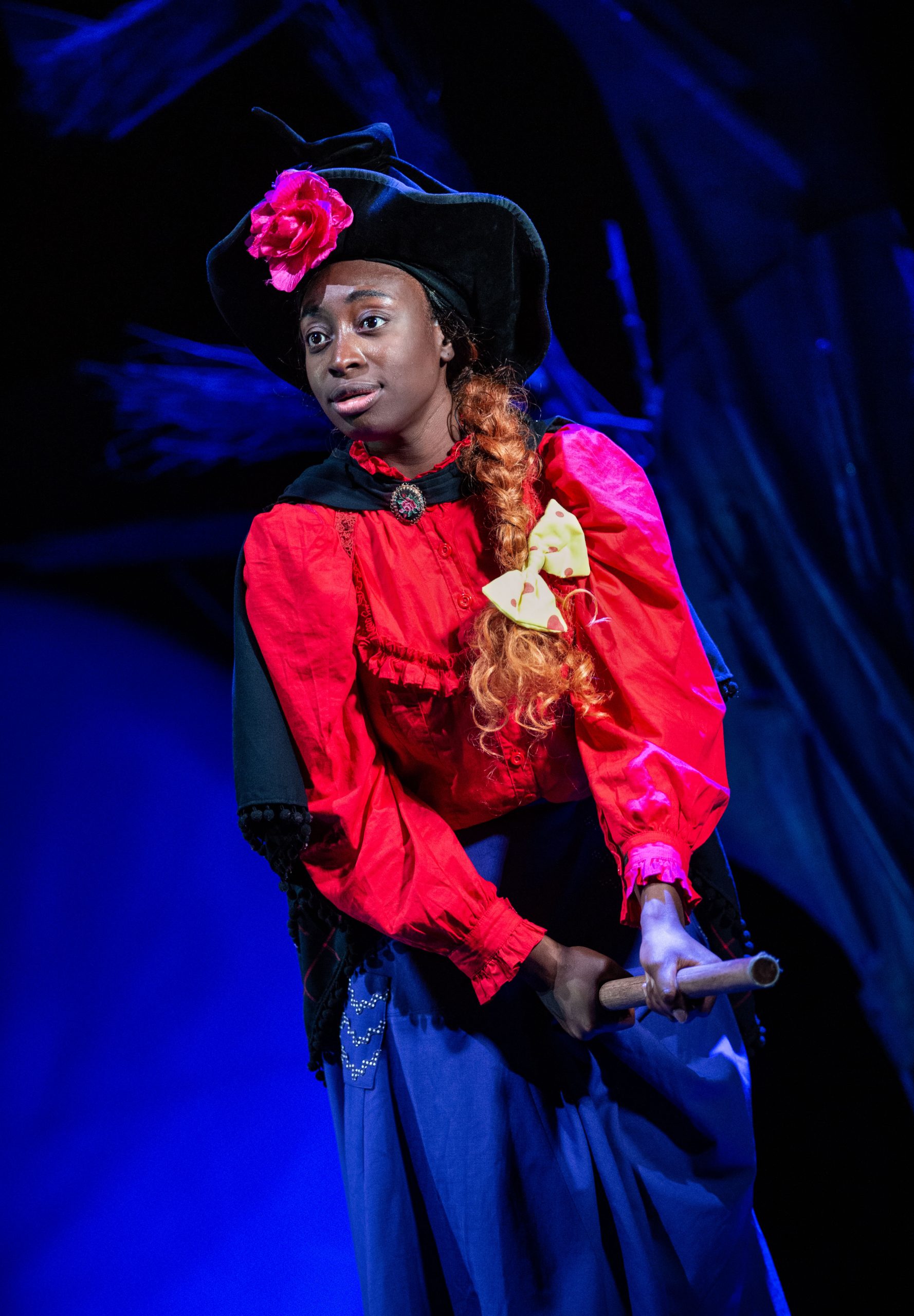 The witch wearing a red shirt and purple skirt. She has orange hair in a long plait with a yellow bow. On top of her head is a long, pointed black witch