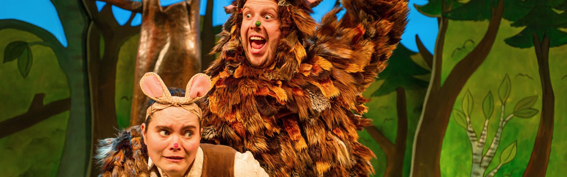 Actor in mouse costume cowering below an actor in Gruffalo costume who joyfully looks like they are about to eat the mouse.