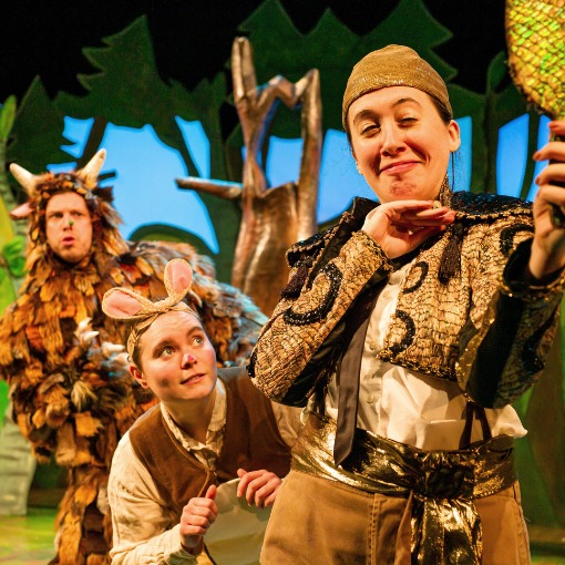 Actor in Snake costume vainly staring into a mirror with actors in Mouse and Gruffalo costume in the background in front of a wood set.