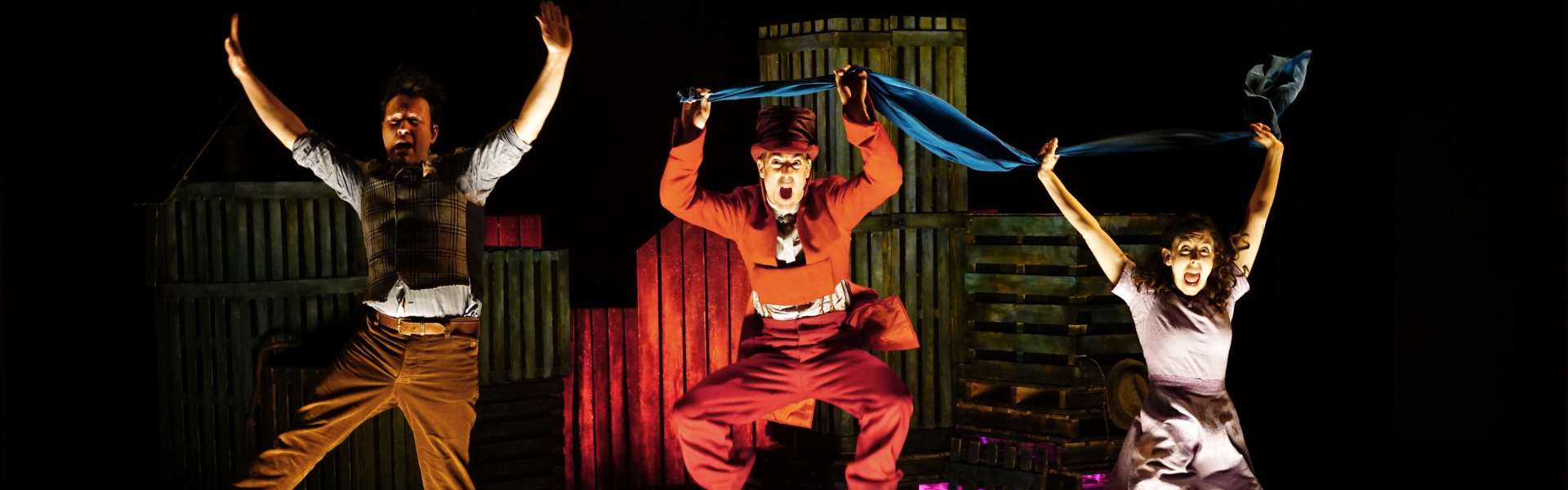 Three people mid-jump in the air. The people in the middle and on the right are holding a blue piece of cloth between them. The woman on the right is wearing a lilac dress. The middle man is wearing a red suit and top hat. The man on the left is wearing a brown/tan trousers and waistcoat with a white shirt.
