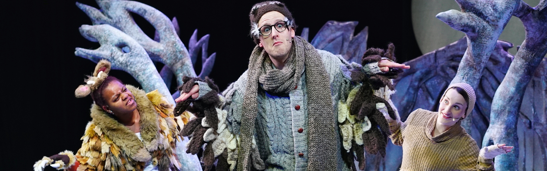 A man in a grey wooly cardign and scarf pretending to be an owl. He is wearing glasses and looking directly at the camera. To his left is a woman dressed as the Gruffalo