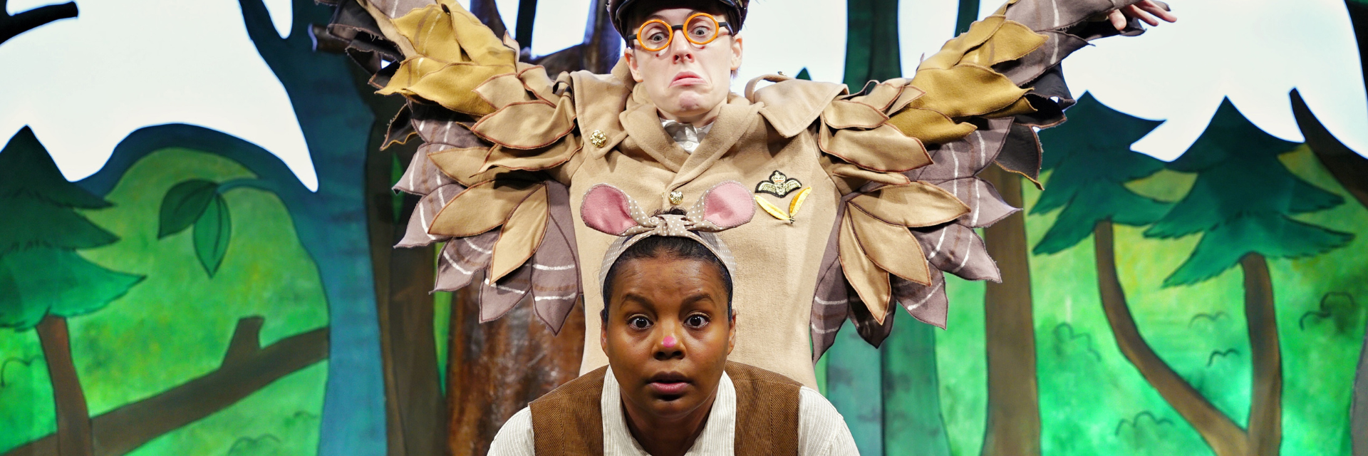 A woman dressed as The Mouse from The Gruffalo dressed in a white top and brown waistcoat. Behind her is a man dressed as an Owl, with his arms raised, ready to pounce.