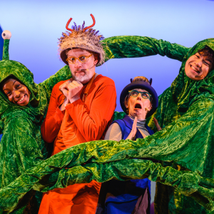 Two actors dressed as Grandfather Smed and Grandmother Smoo from The Smeds and The Smoos. They are trapped between two aliens with long green arms and three eyes on top of their heads. In the background is a blue rocket.