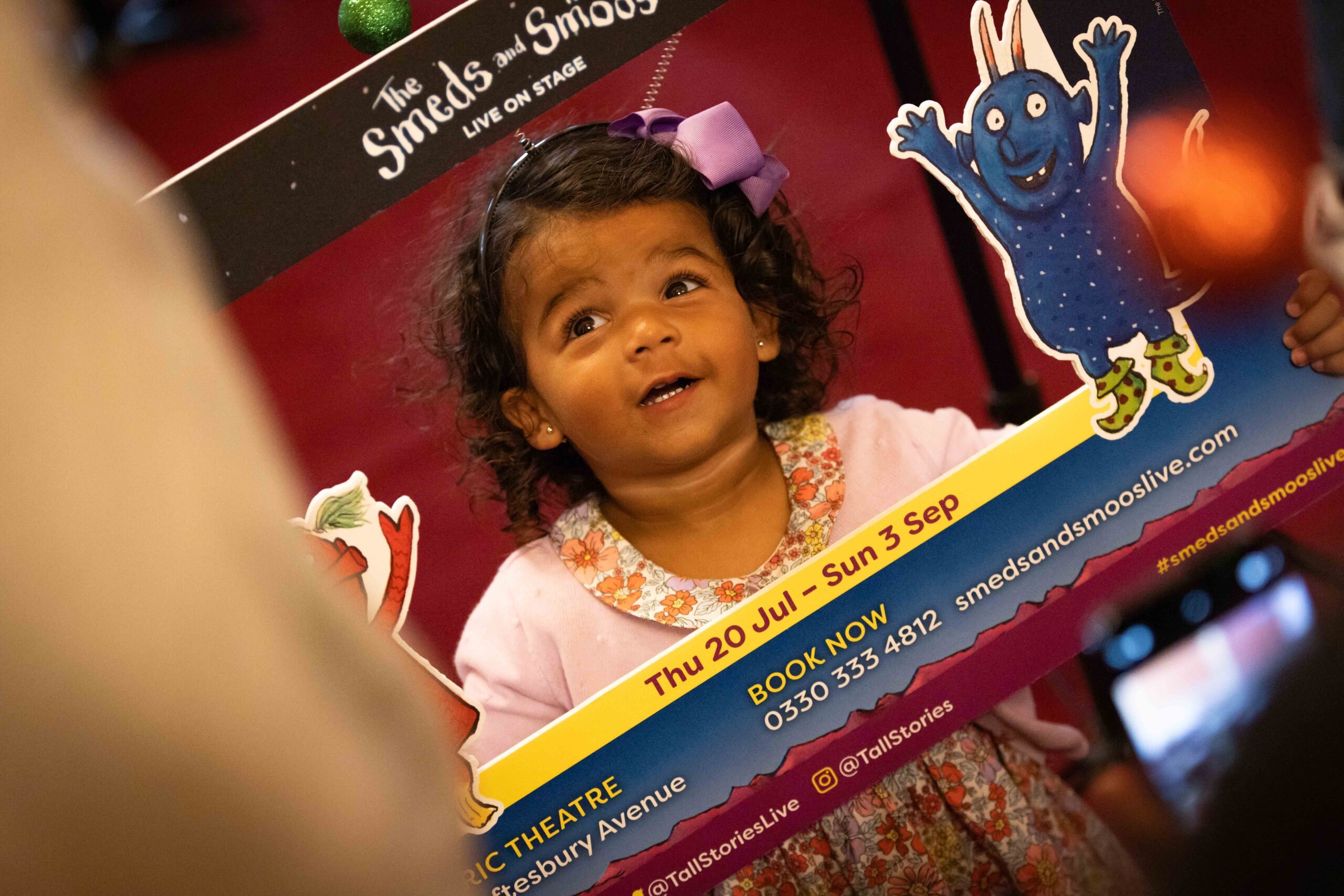 A young girl in a dress and pink cardigan holding up a photo booth frame from The Smeds and The Smoos.