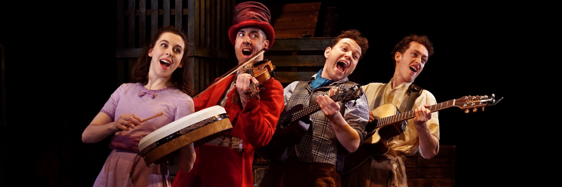 Four performers playing instruments. the woman on the left is in a purple dress and playing a drums. The man next to her is playing a violin and wearing a red suit with red top hat. Next to them are two men in shirts playing guitars. They are all singing.
