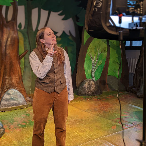 A woman with brown hair wearing a brown waistcoat, white shirt and orange trousers doing sign language. She is performing this to some filming equipment.