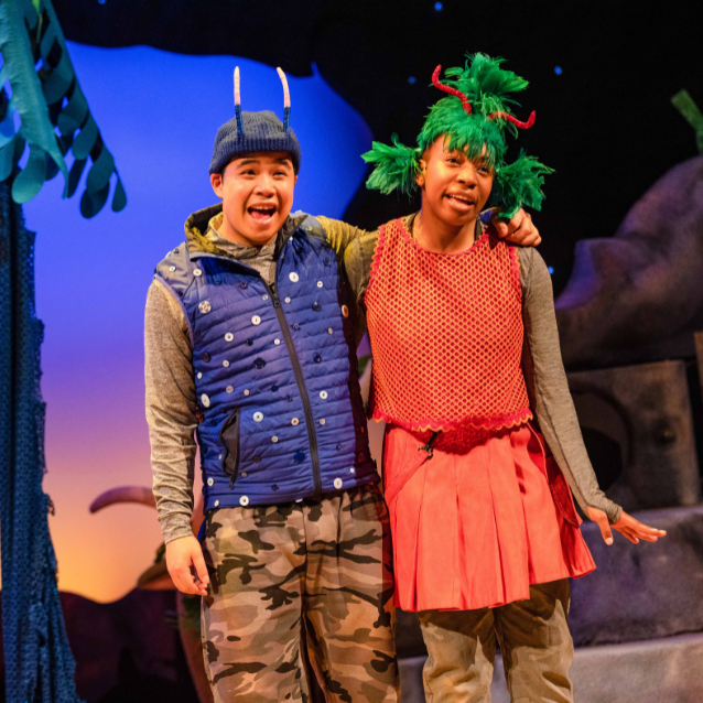An actor playing a blue Smoo with his arm around an actress playing a red Smed. They are both singing.