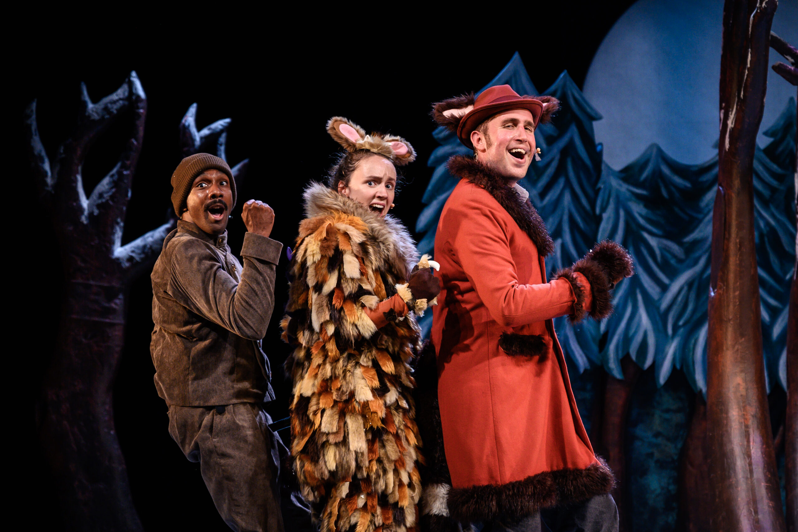 Three actors singing and dancing with the forearms bent. They are in a line. One is wearing a red coat, one a Gruffalo