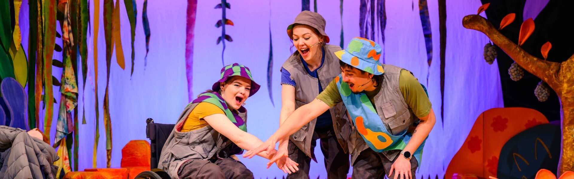 hree actors stand on a colorful stage dressed as campers, wearing bucket hats, vests, and cargo pants. One actor uses a wheelchair and wears a vibrant purple and green sash, while the others cheerfully stand behind her with tie-dye flags and jungle foliage as the backdrop. They all have their left arms pointing into the centre of the circle, with their hands on top of each other.