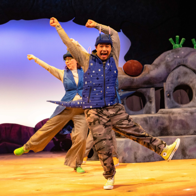 A man in the middle of dancing and looking jubilant. He is wearing a bright blue hat and gilet and brown trouser. There is a woman dressed similarly behind him and also dancing