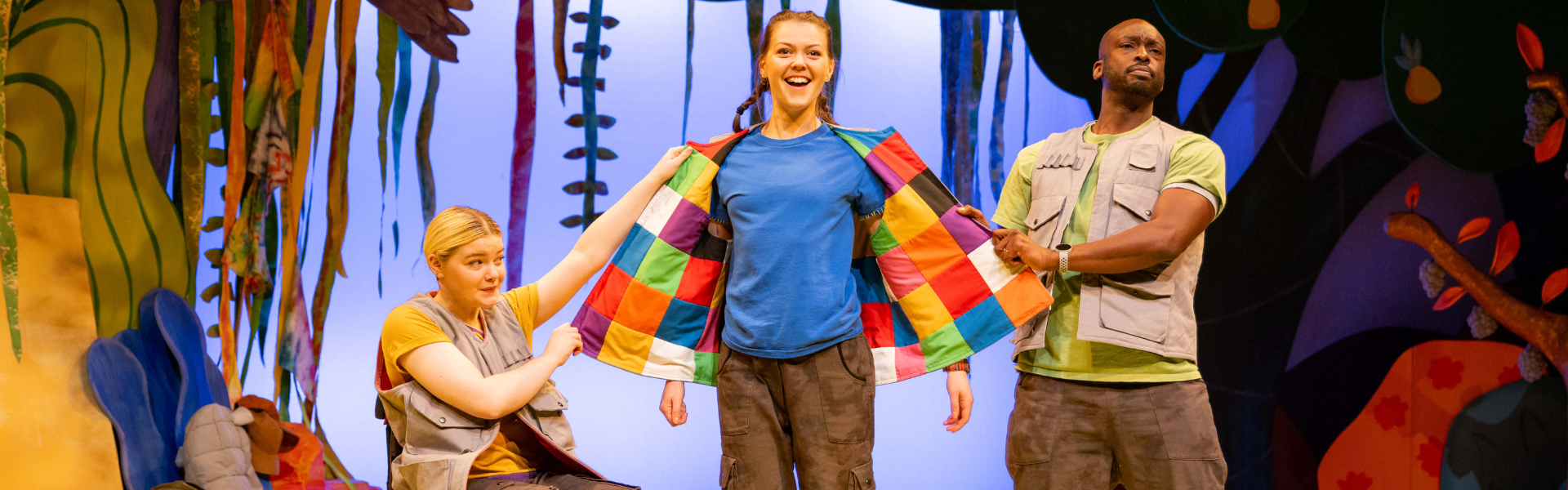 Three actors perform on a colorful stage set resembling a jungle. The central actor, wearing a blue shirt, stands confidently with a wide smile as two others, wearing utility vests, hold out the panels of a patchwork coat they are wearing. The scene is lively, with exaggerated expressions and a whimsical backdrop of oversized plants and vines.