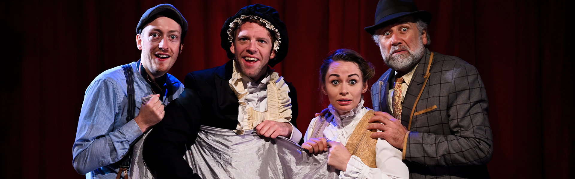 Four actors holding a cloth and facing the camera. They are all in smart Victorian-era clothing and look shocked.