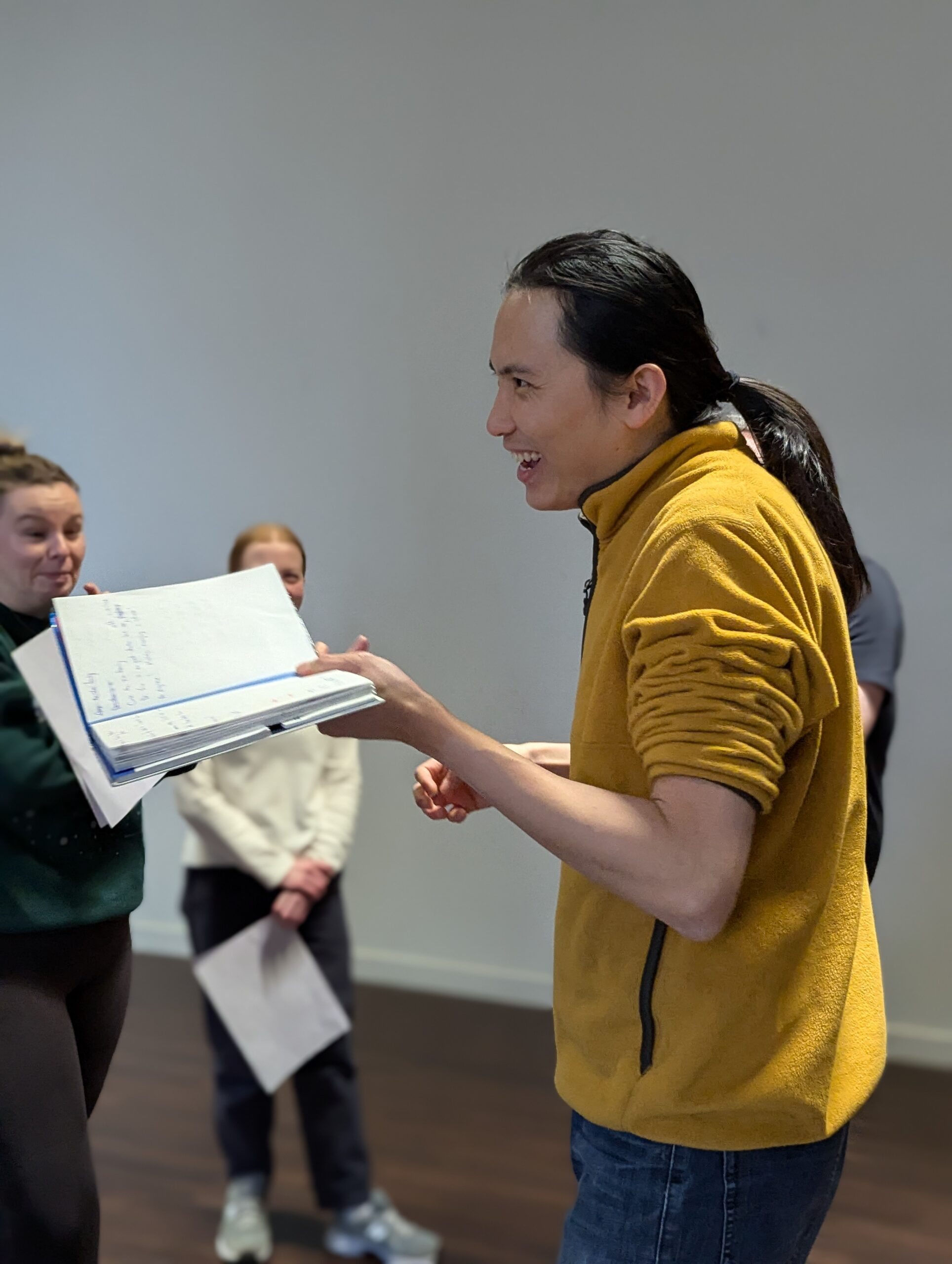 A man with long black hair tied back in a pony tail standing wiht a notebook in his hand wearing a yellow fleece jacket