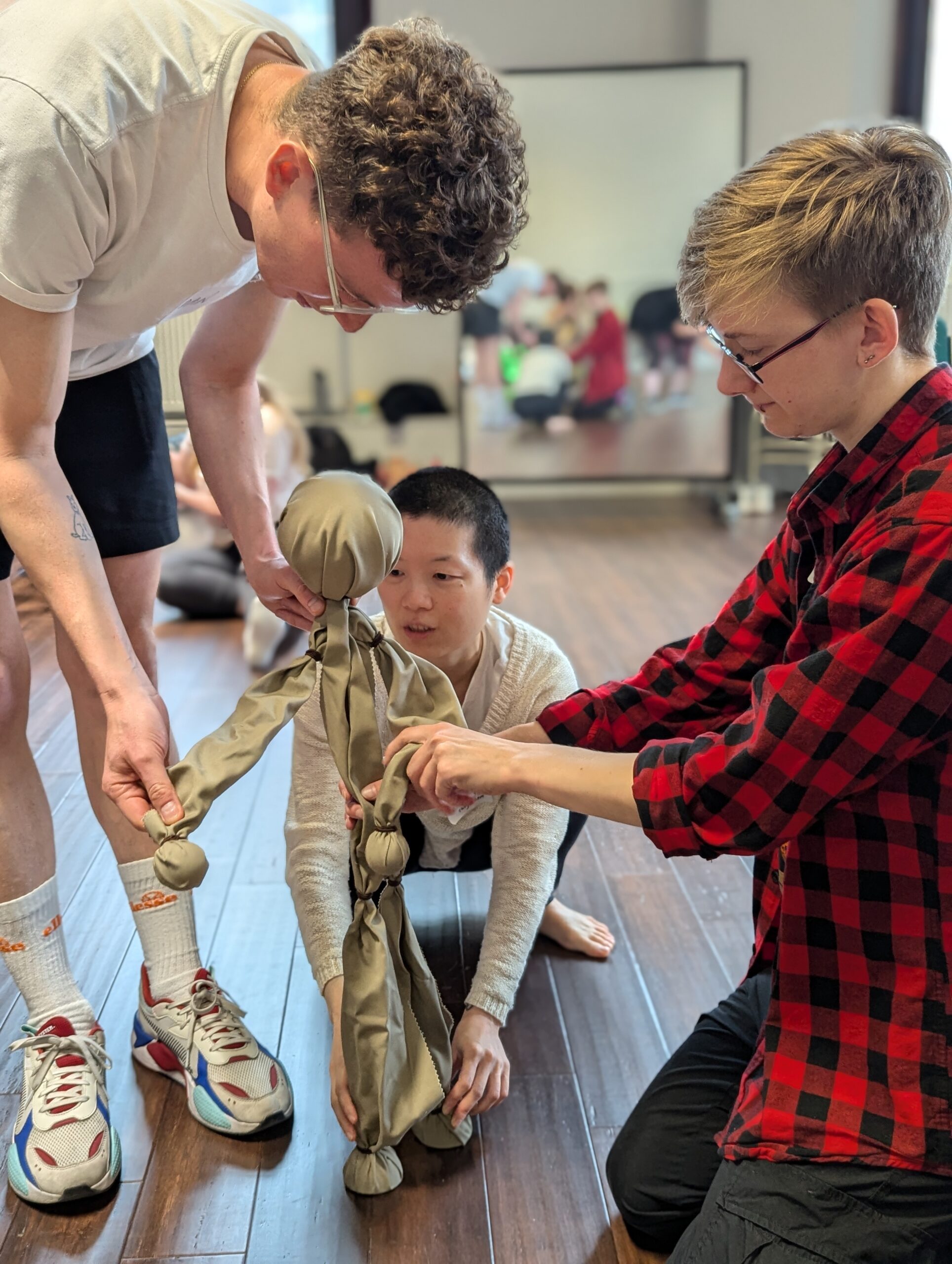 Three people on a studio floor playing with a simple white puppet