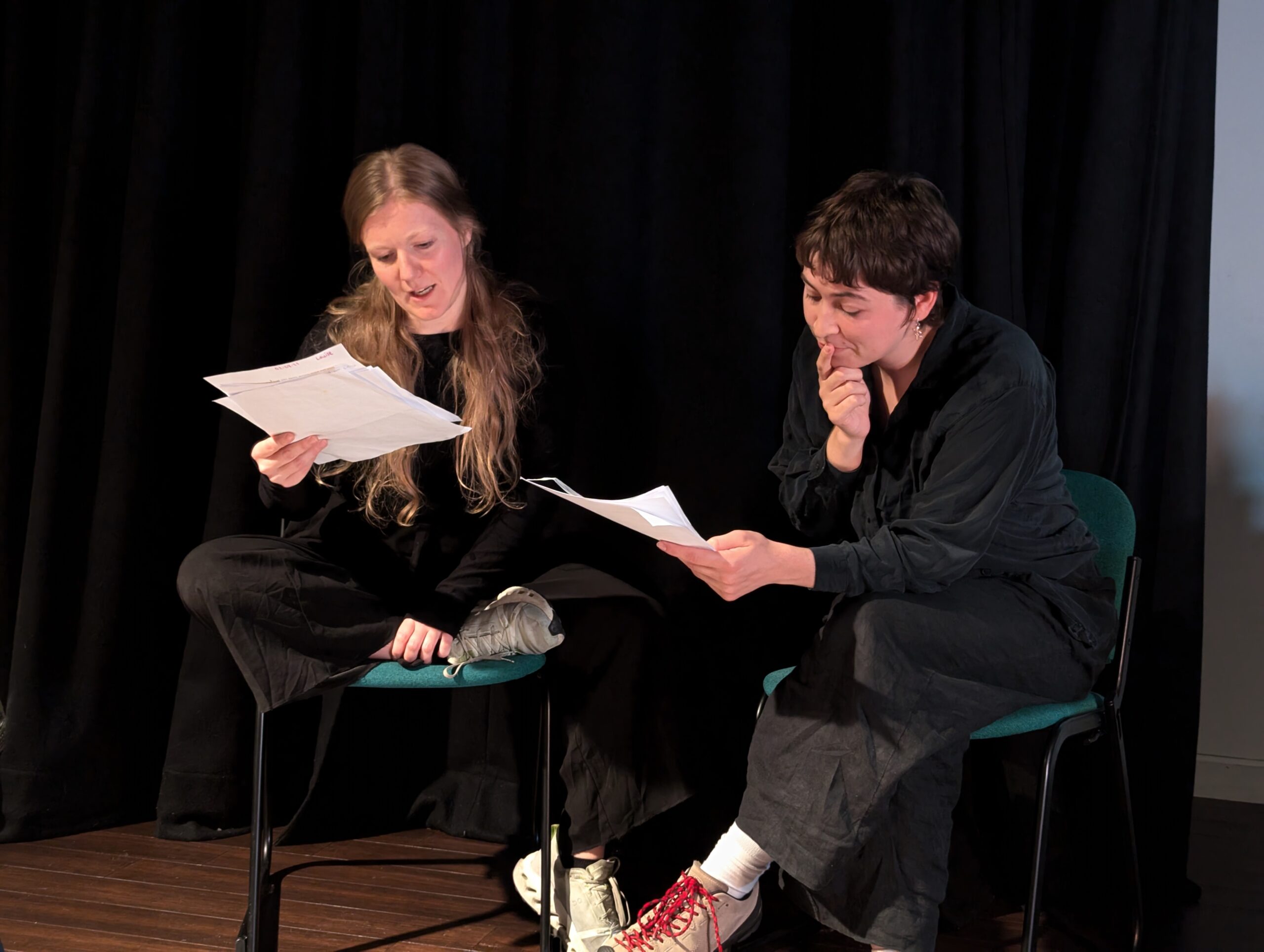 Two people dressed in dark clothing sitting down holding pieces of paper that they are reading