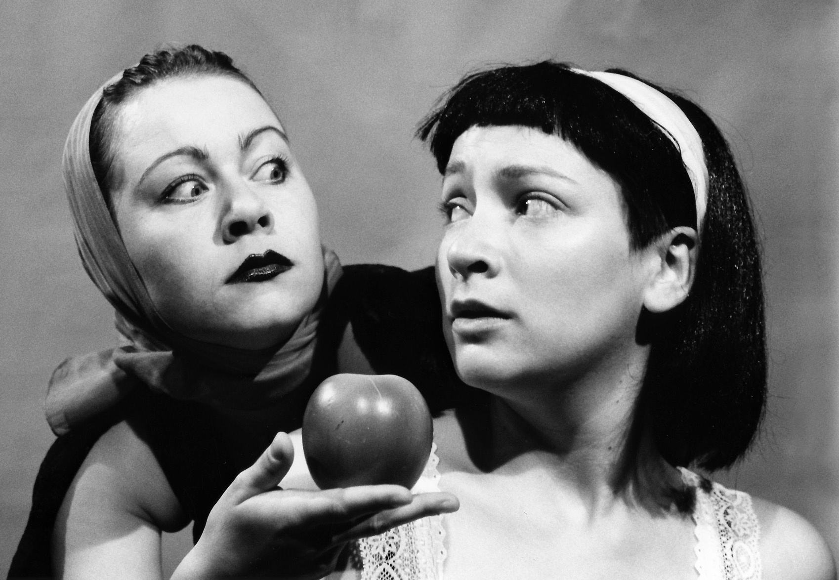 Two women are captured in dramatic grayscale. One, with a headscarf, gazes intensely at the other, who holds an apple, conveying tension and curiosity.