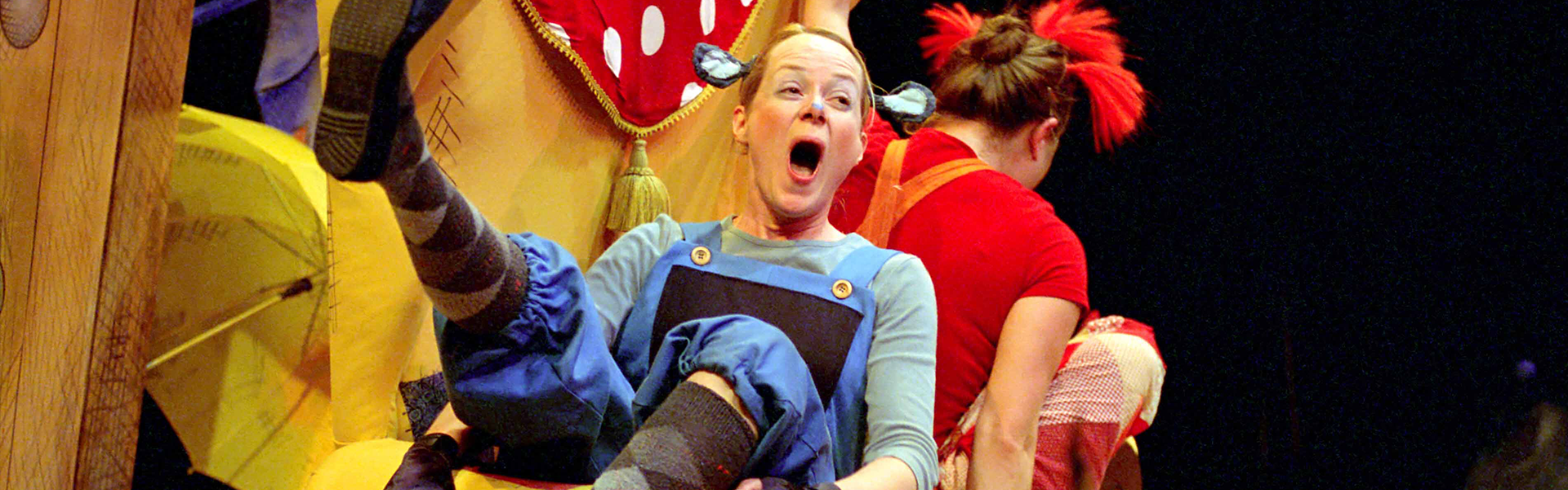 A joyful performer in blue overalls with blue ears and striped socks playfully kicks a leg high on an oversized chair. Another performer in red sits on the other side of the chair with their back to the camera. A whimsical atmosphere with bright, eclectic outfits.