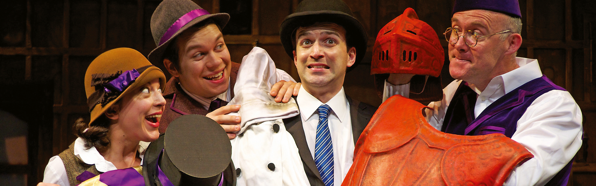 Four actors in colourful costumes share humorous expressions on stage. One holds a red helmet, creating a playful and comedic tone.