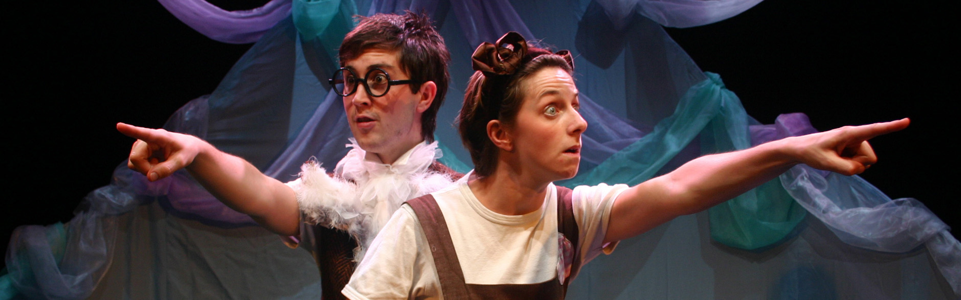 Two actors on stage in whimsical costumes, dressed as an owl and cat, point dramatically in different directions. The background features colourful, flowing fabric, adding a playful tone.