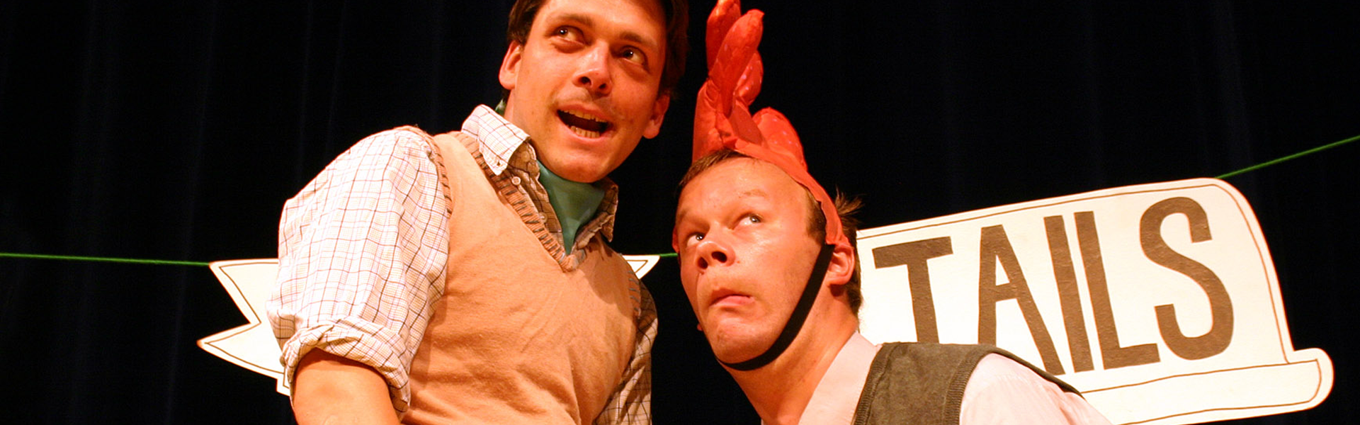 Two actors perform on stage, one wearing a rooster hat with a playful expression. A sign with the word 